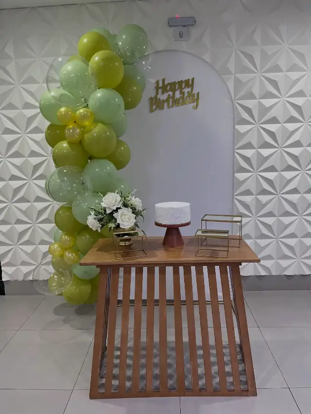 Mesa de madeira com bolo branco, arco de balões verdes e painel em arco com Happy Birthday